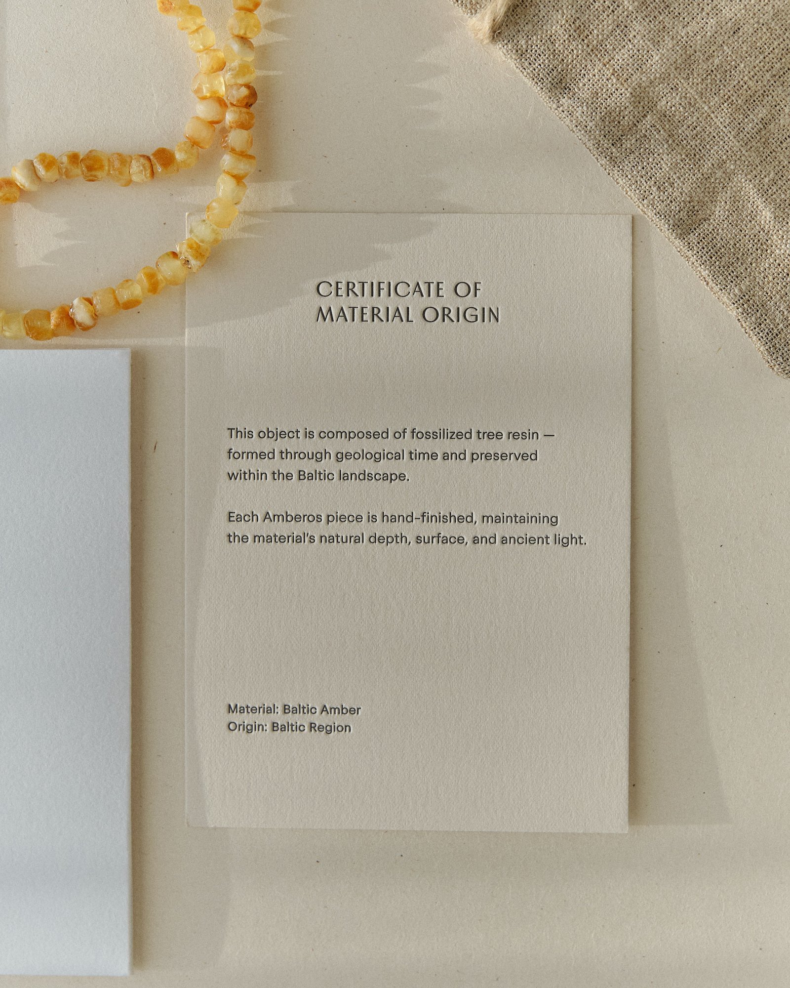 Pale amber necklace beside the Amberos certificate of material origin card and packaging box on linen