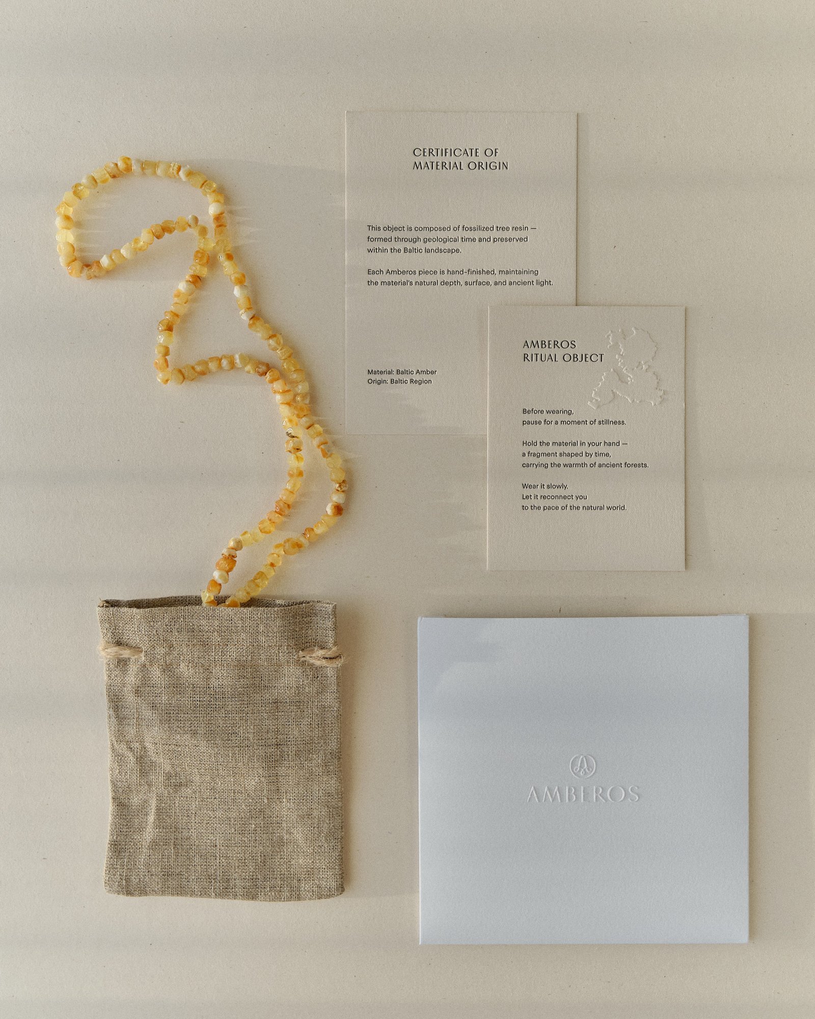 Pale amber necklace laid across the Amberos certificate card, ritual object card, linen pouch and box, seen from above