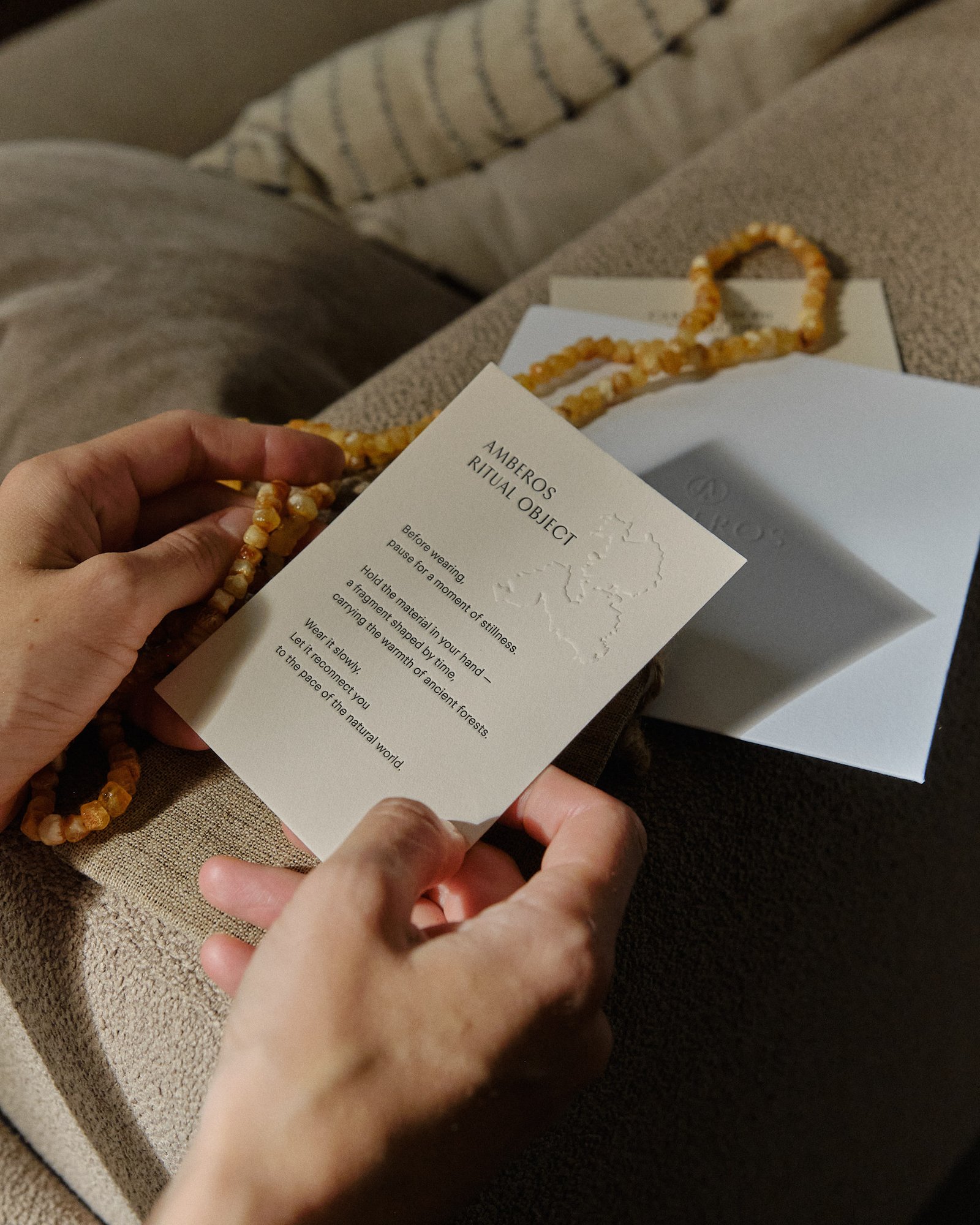 Hands holding the Amberos ritual object card with a honey amber necklace draped over a linen pouch