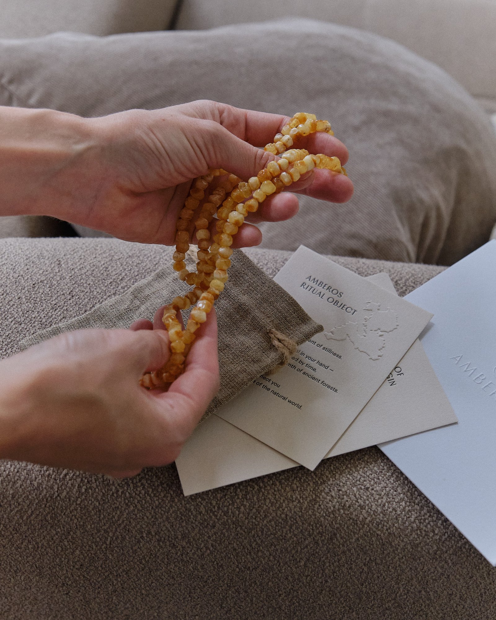 Hands holding a pale amber necklace above the Amberos packaging cards on a sofa