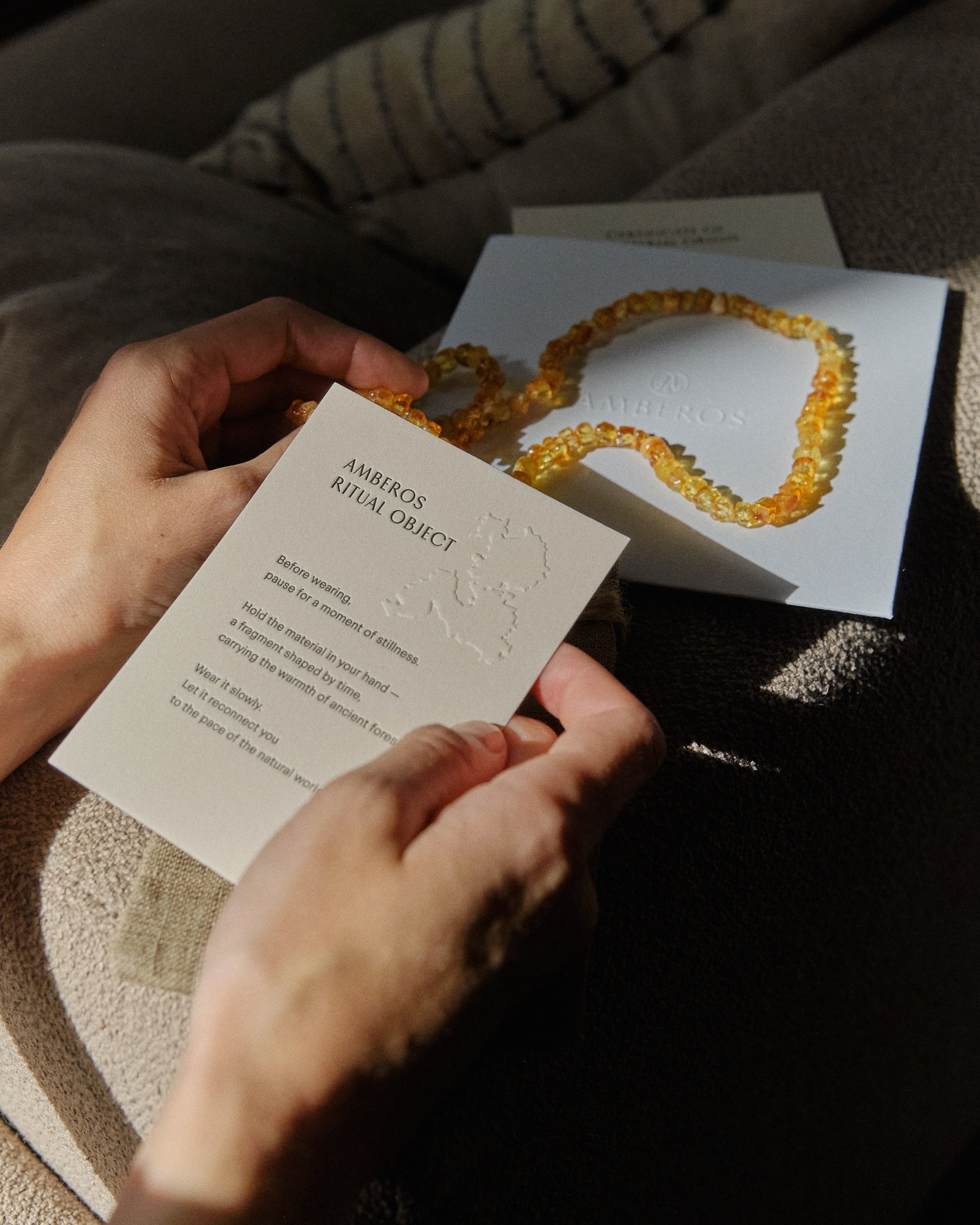Hands holding the Amberos ritual object card and certificate card with a cognac amber necklace in dim evening light