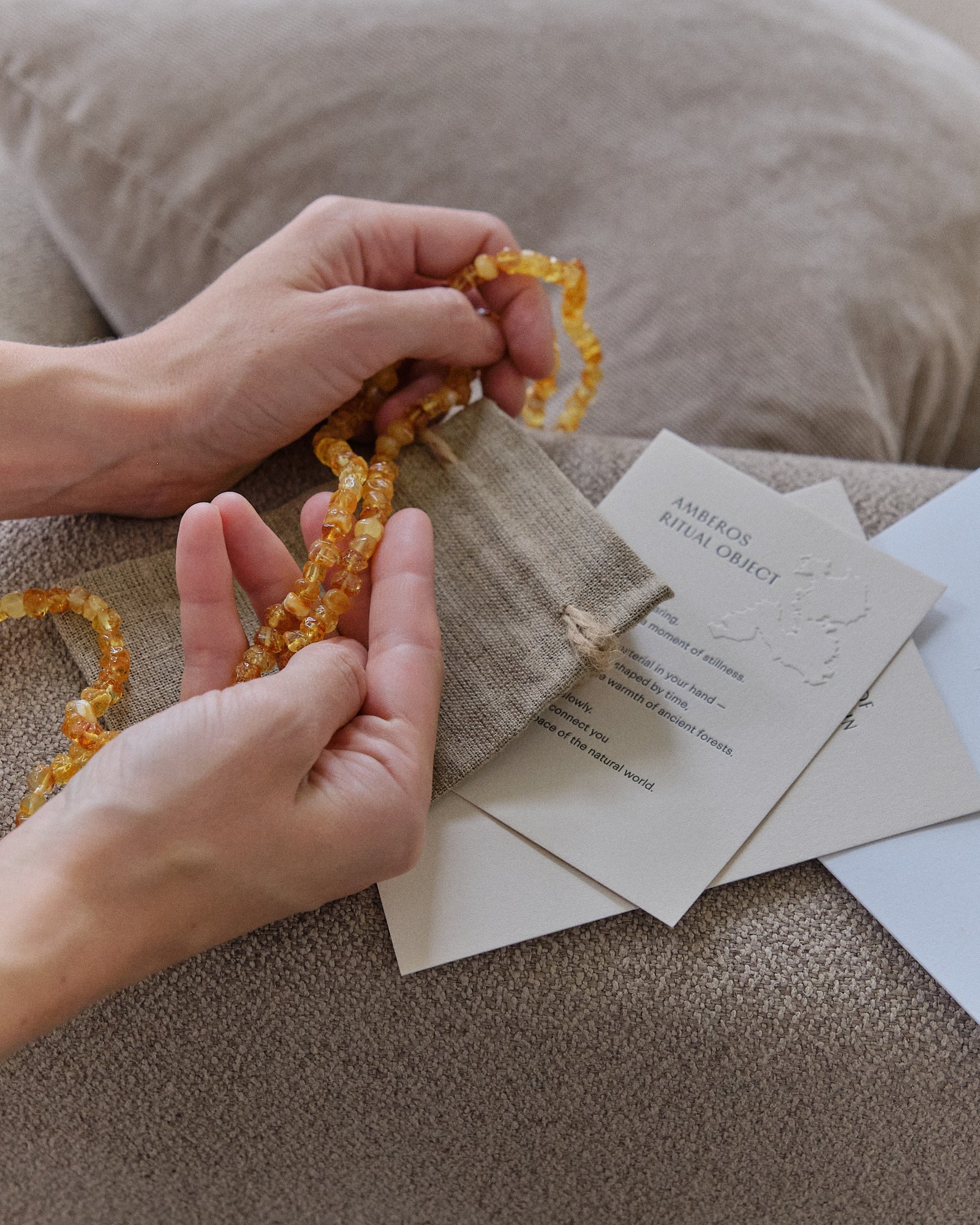 Hands holding a honey amber necklace above the Amberos linen pouch and ritual object card on a sofa