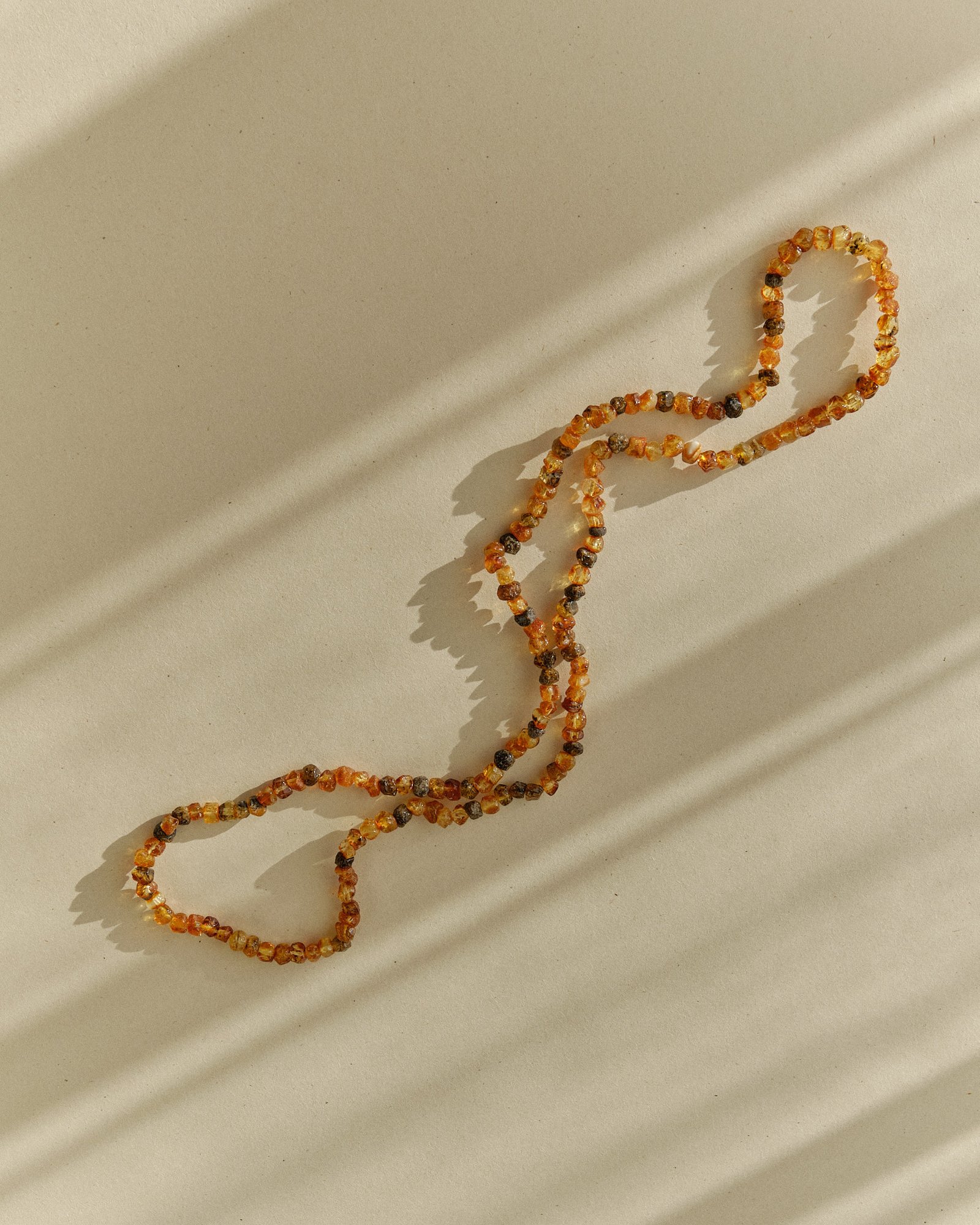 Close-up of a cognac and dark amber necklace winding in an S shape on a sunlit surface with long streaked shadows