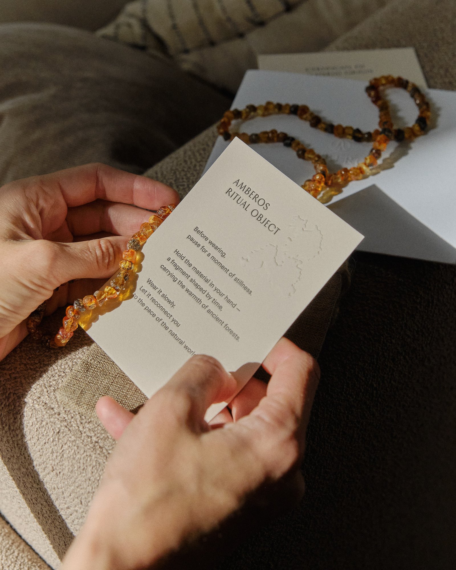 Hands holding the Amberos ritual object card with a cognac amber necklace on a sofa