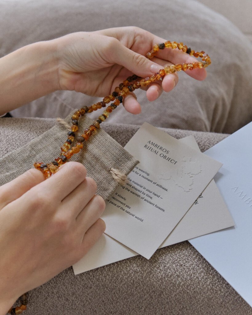 Hands holding a cognac and dark amber necklace beside the Amberos ritual object card on a sofa